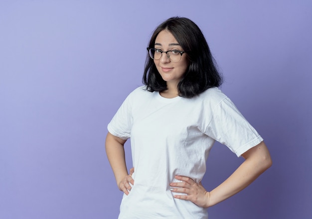 Pleased young pretty caucasian girl wearing glasses putting hands on waist isolated on purple background with copy space