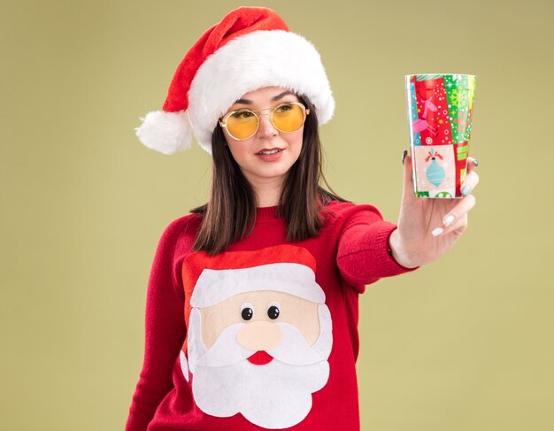올리브 녹색 배경에 고립 된 그것을보고 카메라를 향해 플라스틱 크리스마스 컵을 뻗어 안경 산타 클로스 스웨터와 모자를 쓰고 기쁘게 젊은 예쁜 백인 여자