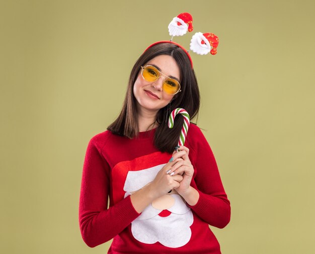 コピースペースでオリーブグリーンの背景に分離されたカメラを見て伝統的なクリスマスのキャンディケインを保持しているメガネとサンタクロースのセーターとヘッドバンドを身に着けている若いかなり白人の女の子を喜ばせる
