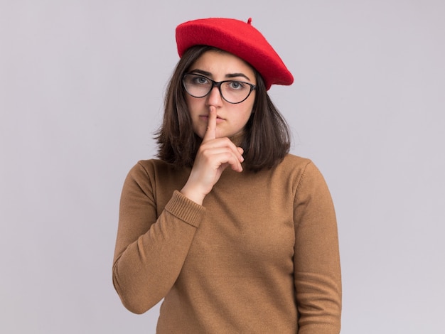 Pleased young pretty caucasian girl with beret hat and in optical glasses doing silence gesture isolated on white wall with copy space