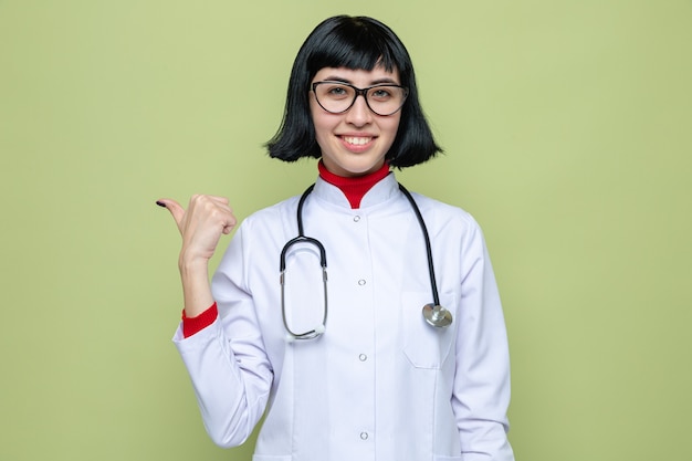 Lieta giovane bella donna caucasica con gli occhiali in uniforme da medico con uno stetoscopio rivolto a lato