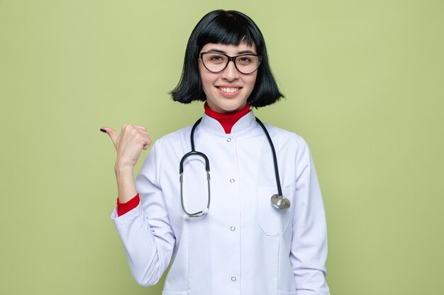 Довольная молодая симпатичная кавказская женщина в очках в униформе врача со стетоскопом, указывающим в сторону