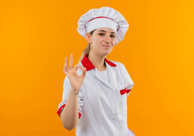 Pleased young pretty cook in chef uniform doing ok sign looking  isolated on orange space