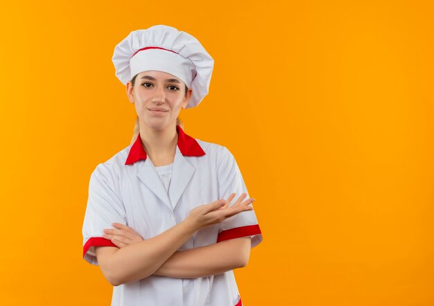 閉じた姿勢で立って、オレンジ色のスペースで隔離された空の手を示すシェフの制服を着た若いかわいい料理人を喜ばせます