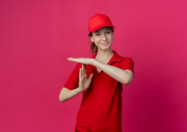 Pleased young pretty delivery girl in red uniform and cap doing timeout gesture isolated on crimson background with copy space