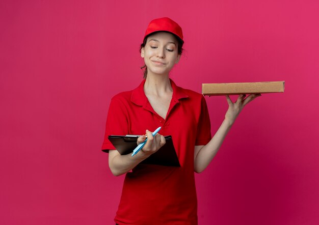 빨간 제복을 입은 만족 된 젊은 예쁜 배달 소녀와 모자 복사 공간이 진홍색 배경에 고립 된 클립 보드를보고 피자 패키지와 클립 보드와 펜을 들고 모자