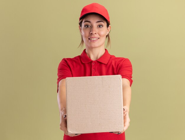 Pleased young pretty delivery girl in uniform holds cardbox on olive green