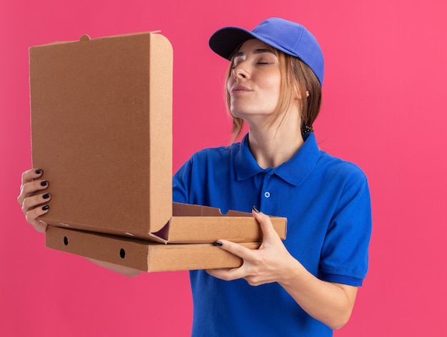 Soddisfatta la giovane e graziosa ragazza delle consegne in uniforme tiene le scatole della pizza e finge di annusare il rosa