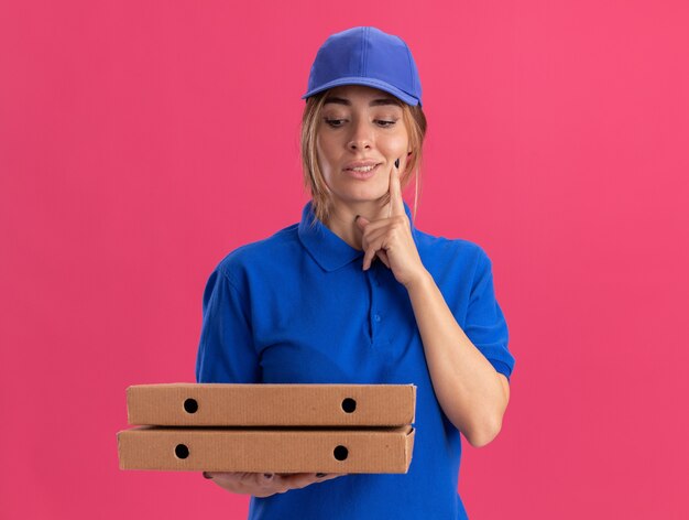 Pleased young pretty delivery girl in uniform puts finger on face holding and looking at pizza boxes on pink