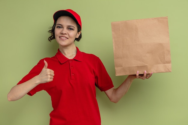 Pleased young pretty delivery woman holding paper food packaging and thumbing up