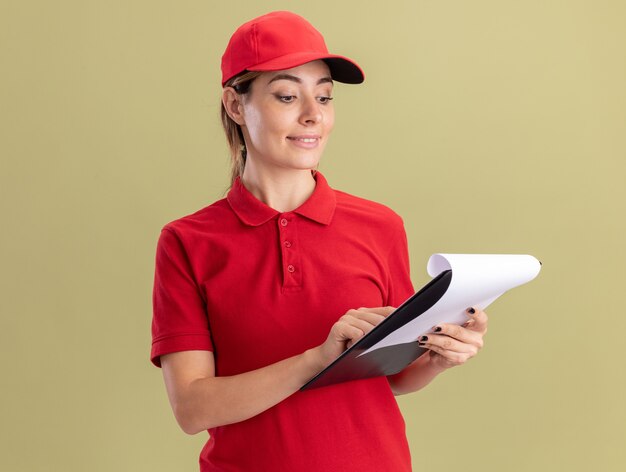 Pleased young pretty delivery woman in uniform holds and looks at clipboard isolated on olive green wall