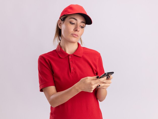 Pleased young pretty delivery woman in uniform holds and looks at phone isolated on white wall