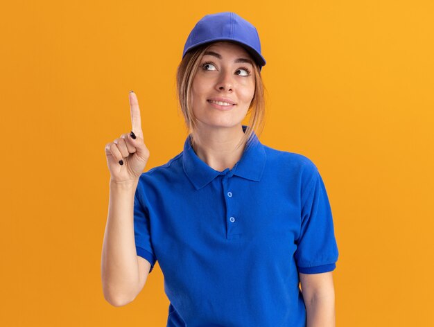 Pleased young pretty delivery woman in uniform points up isolated on orange wall