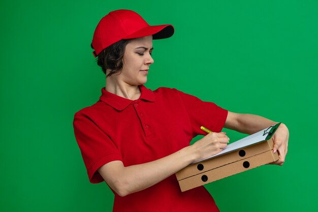 피자 상자를 들고 클립보드에 글을 쓰는 행복한 젊은 예쁜 배달부