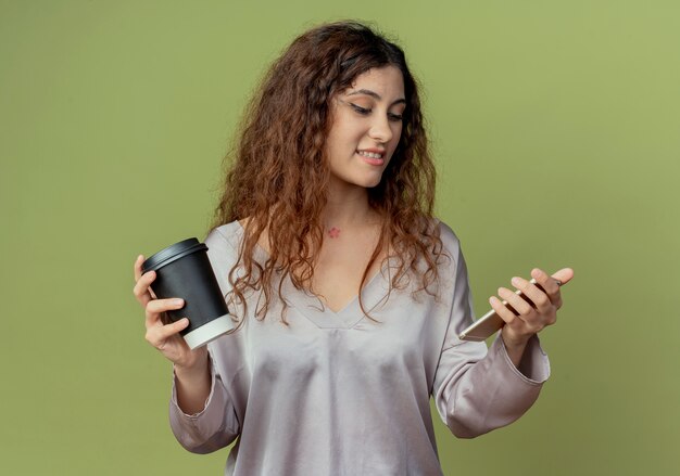 Pleased young pretty female office worker holding cup of coffee and looking at phone in her hand isolated on olive green wall