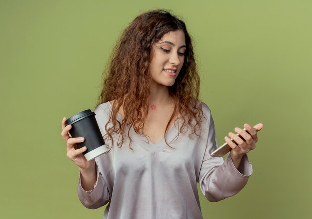 Довольный молодой симпатичный женский офисный работник, держащий чашку кофе и смотрящий на телефон в руке, изолированной на оливково-зеленой стене