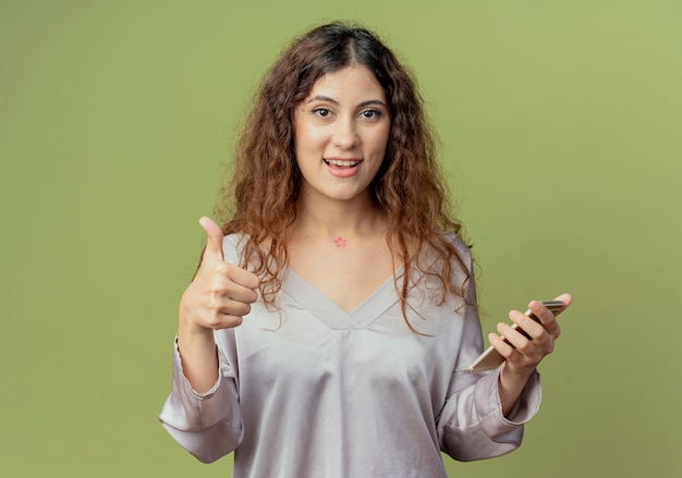 Pleased young pretty female office worker holding phone and her thumb up isolated on olive green wall