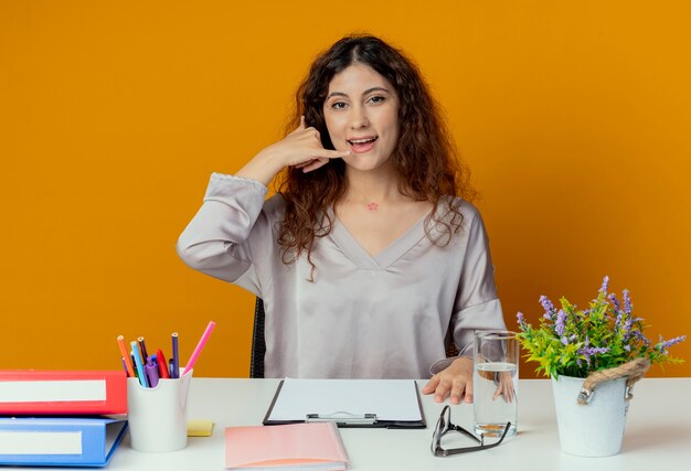 オレンジ色に分離された電話ジェスチャーを示すオフィスツールで机に座っている若いきれいな女性のサラリーマンを喜ばせる