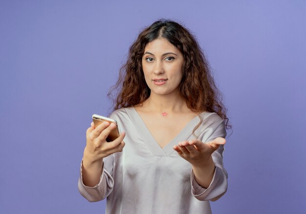 Pleased young pretty girl holding phone and holding out hand to camera isolated on blue