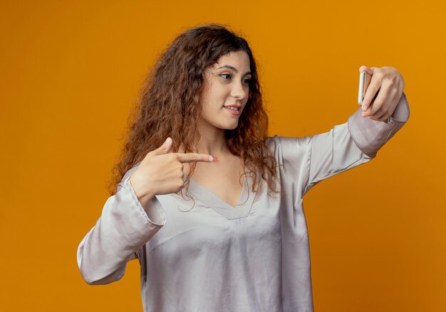 Pleased young pretty girl take a selfie and points at phone isolated on yellow wall