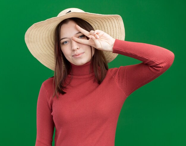 Pleased young pretty girl wearing beach hat showing v-sign symbol near eye 