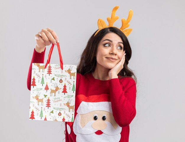 Pleased young pretty girl wearing reindeer antlers headband and santa claus sweater holding christmas gift bag keeping hand on face looking at side 