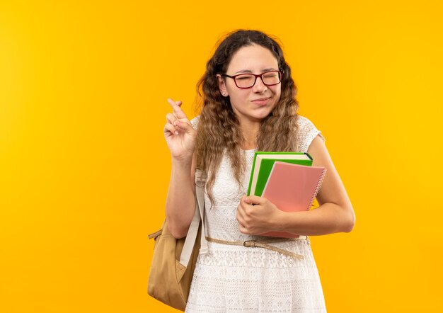 Pleased young pretty schoolgirl wearing glasses and back bag holding book and note pad crossing fingers with closed eyes isolated on yellow wall