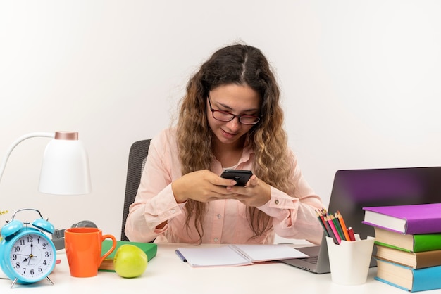 Pleased young pretty schoolgirl wearing glasses sitting at desk with school tools doing her homework using her phone isolated on white wall