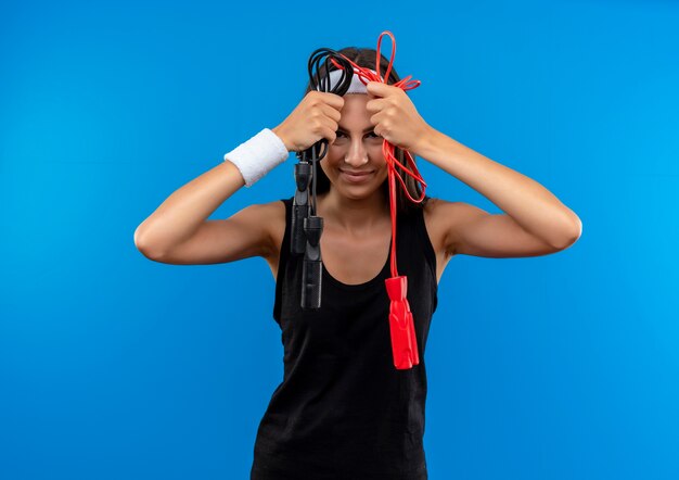 Pleased young pretty sporty girl wearing headband and wristband keeping jumping ropes near head isolated on blue space