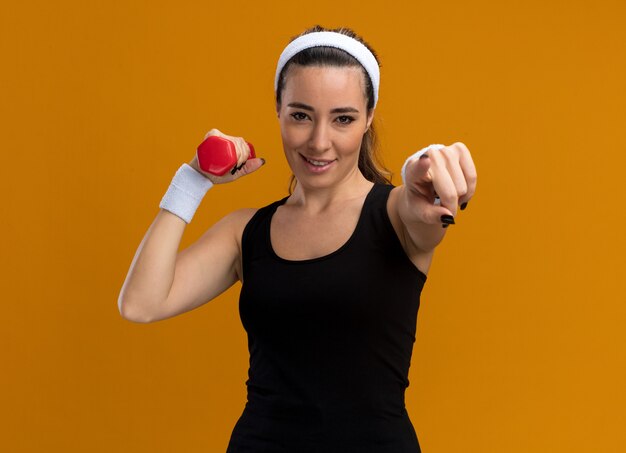 Pleased young pretty sporty girl wearing headband and wristbands holding dumbbell looking and pointing at camera 