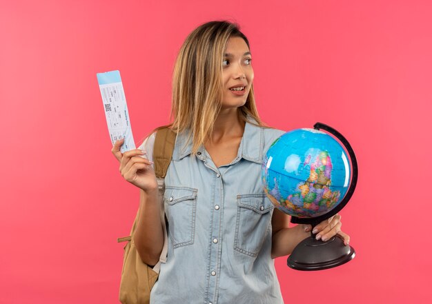 Довольная молодая симпатичная студентка в задней сумке, держащая билет на самолет и глобус, смотрящая в сторону, изолированную на розовой стене