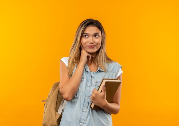 Довольная молодая симпатичная студентка в задней сумке, держащая книгу, кладет руку под подбородок и смотрит в сторону, изолированную на оранжевой стене