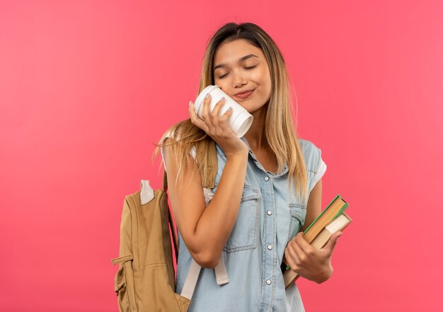 Довольная молодая симпатичная студентка в задней сумке держит книги и трогает лицо пластиковой кофейной чашкой с закрытыми глазами, изолированными на розовой стене с закрытыми глазами