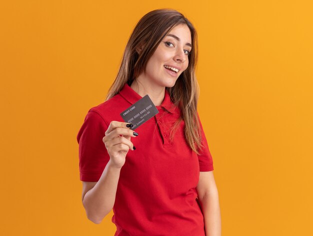 Довольная молодая красивая женщина держит кредитную карту, изолированную на оранжевой стене