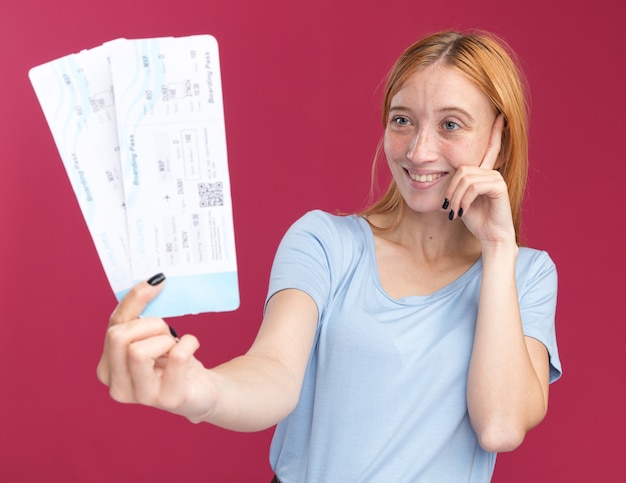 Pleased young redhead ginger girl with freckles puts finger on temple and holds air tickets isolated on pink wall with copy space