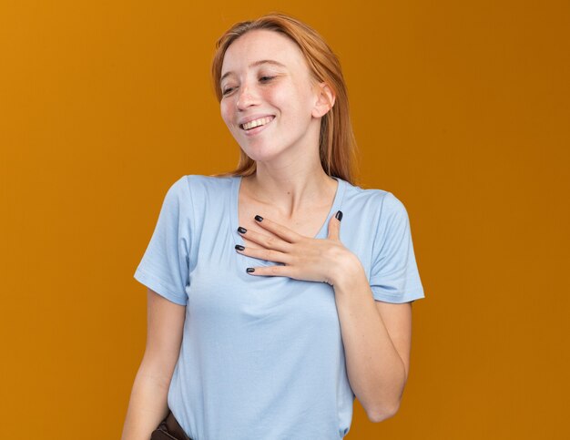 Pleased young redhead ginger girl with freckles puts hand on chest and looks at side isolated on orange wall with copy space