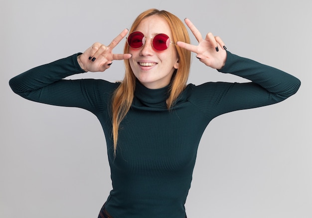 Pleased young redhead ginger girl with freckles in sun glasses gesturing victory sign with two hands on white