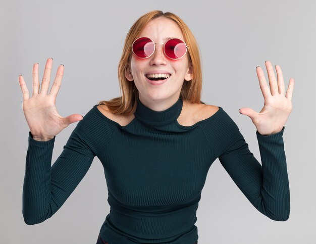 Pleased young redhead ginger girl with freckles in sun glasses standing with raised hands on white