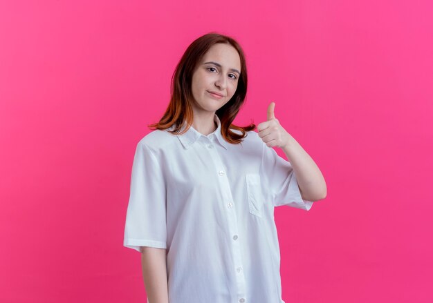 Pleased young redhead girl her thumb up isolated on pink background