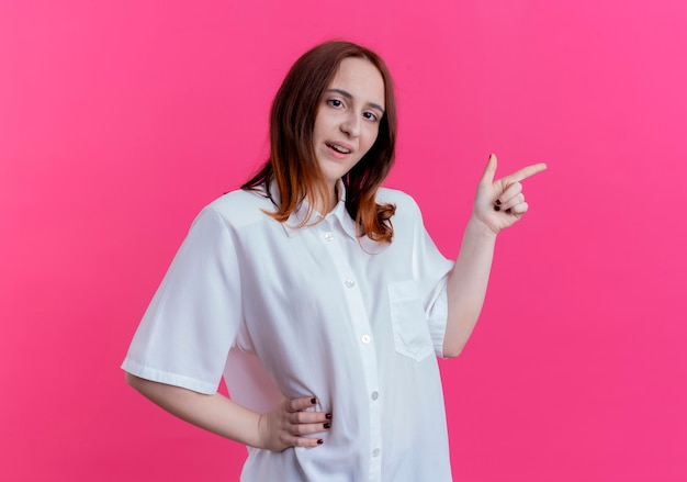 Pleased young redhead girl holding hand on hip and points at side isolated on pink with copy space