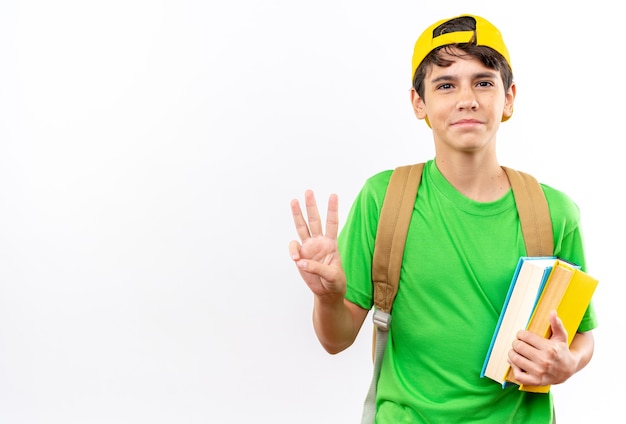 Pleased young school boy wearing backpack with cap holding books showing three isolated on white wall with copy space