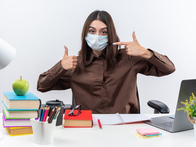 Pleased young school woman wearing medical mask sits at table with school tools showing thumb up 