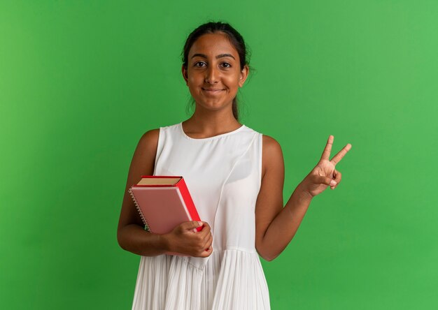 Довольная молодая школьница держит книгу с ноутбуком и показывает жест мира на зеленом