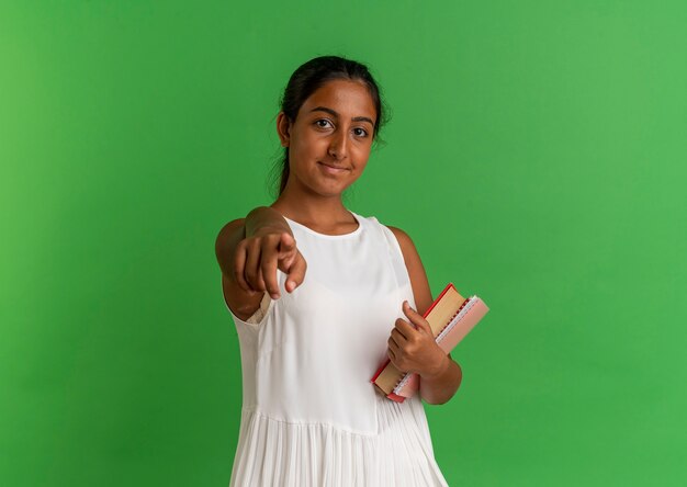 Pleased young schoolgirl holding notebook with book and showing you gesture on green