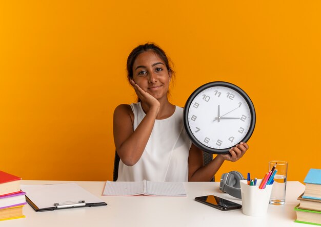 Pleased young schoolgirl sitting at desk with school tools holding wall clock and putting hand on cheek on orange