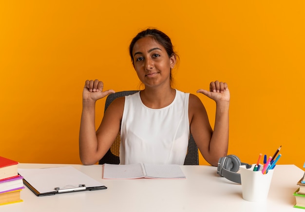 Pleased young schoolgirl sitting at desk with school tools points to herself on orange