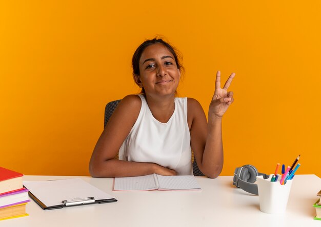 Pleased young schoolgirl sitting at desk with school tools showing peace gesture on orange