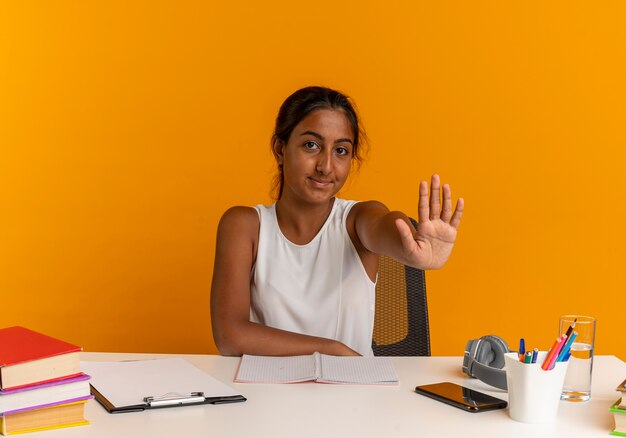 Pleased young schoolgirl sitting at desk with school tools showing stop gesture on orange