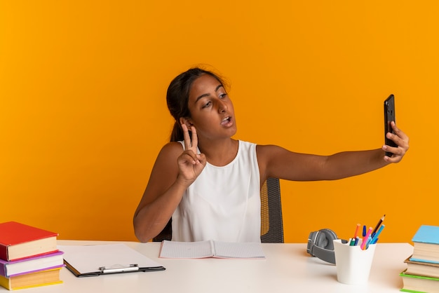 Pleased young schoolgirl sitting at desk with school tools take selfie and showing peace gesture isolated on orange wall