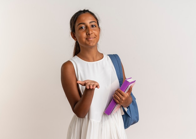 Pleased young schoolgirl wearing back bag holding book and holding out hand isolated on white wall